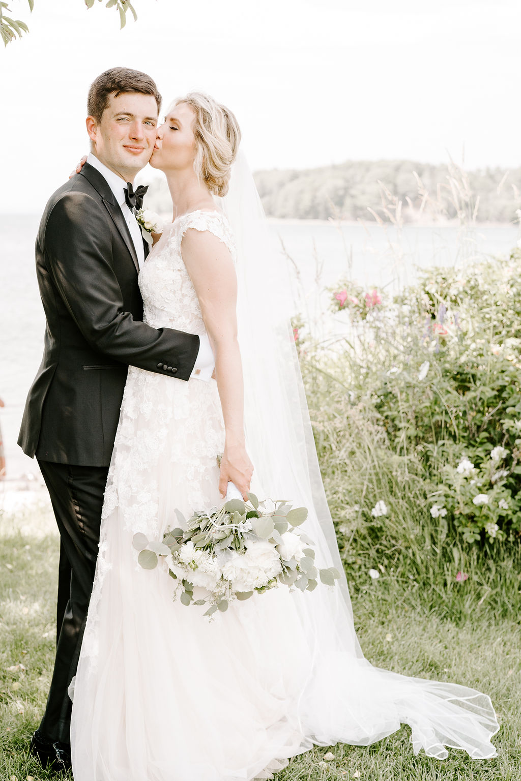 bride kissing groom's cheek on grass overlooking ocean