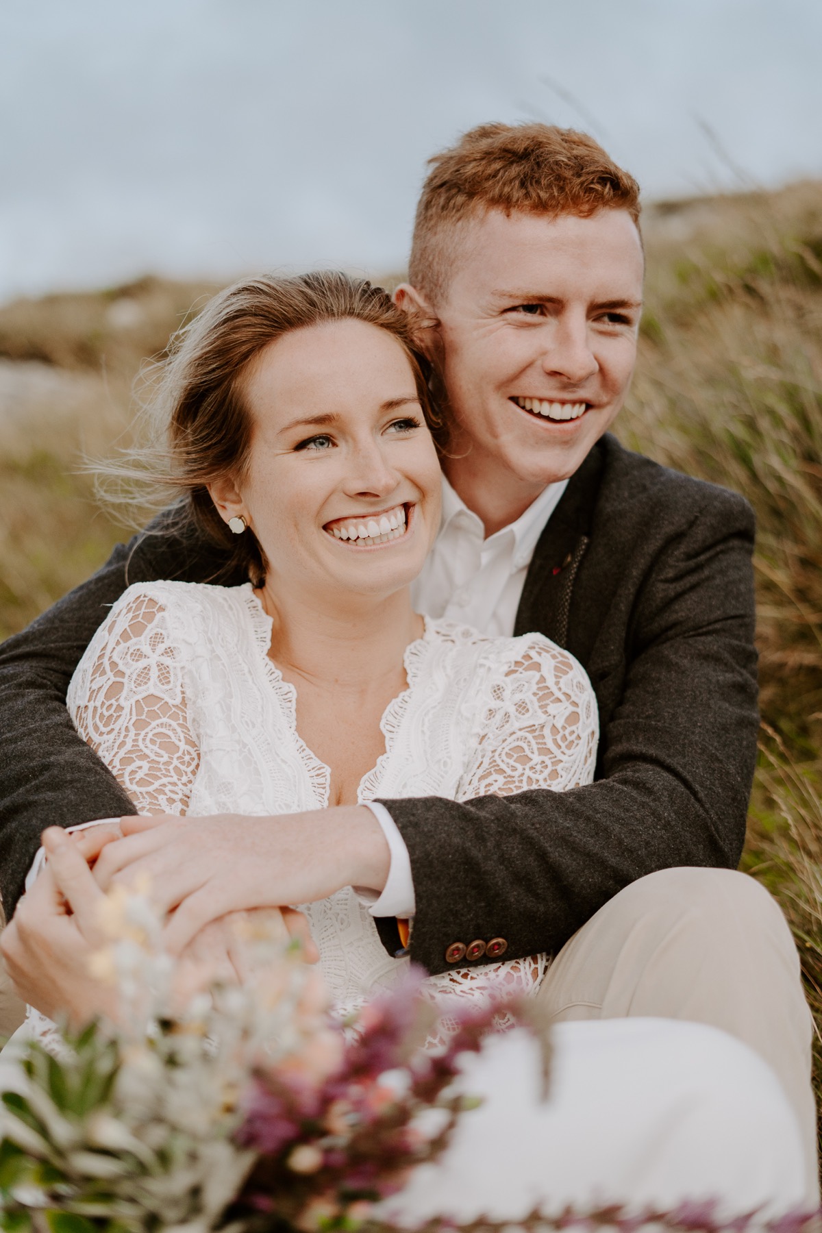 Bride and groom sit together in grass and smile