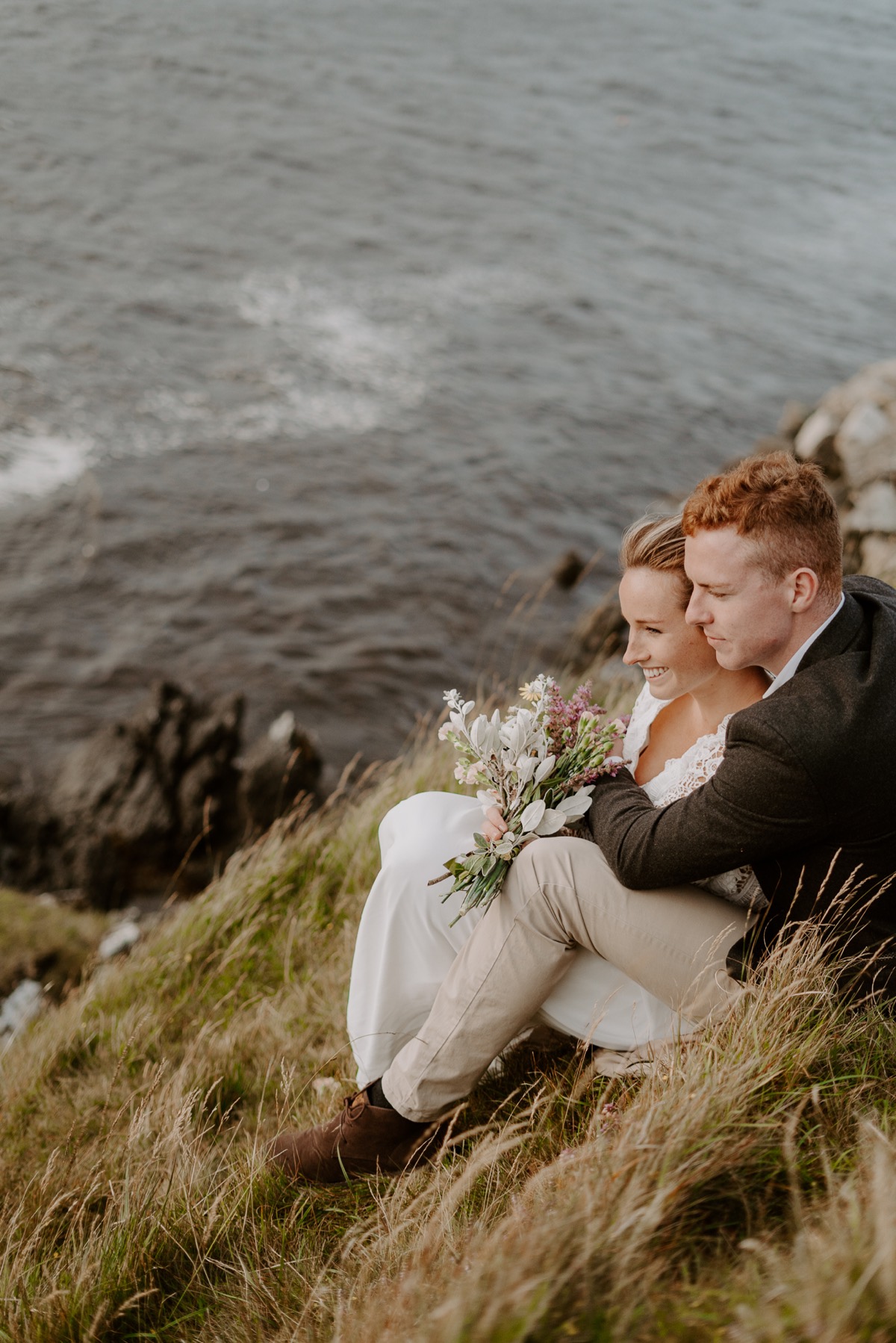 bride and groom sit and look out at ocean from cliff